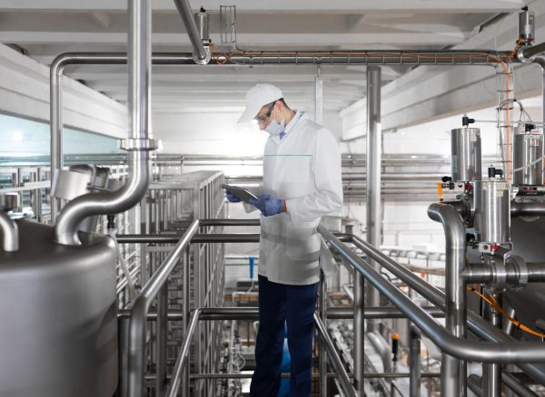 Technologist in a white robe, gloves and mask holding a tablet and stands near the bedon factory dairy products. The inspector checks the performance at the production plant
